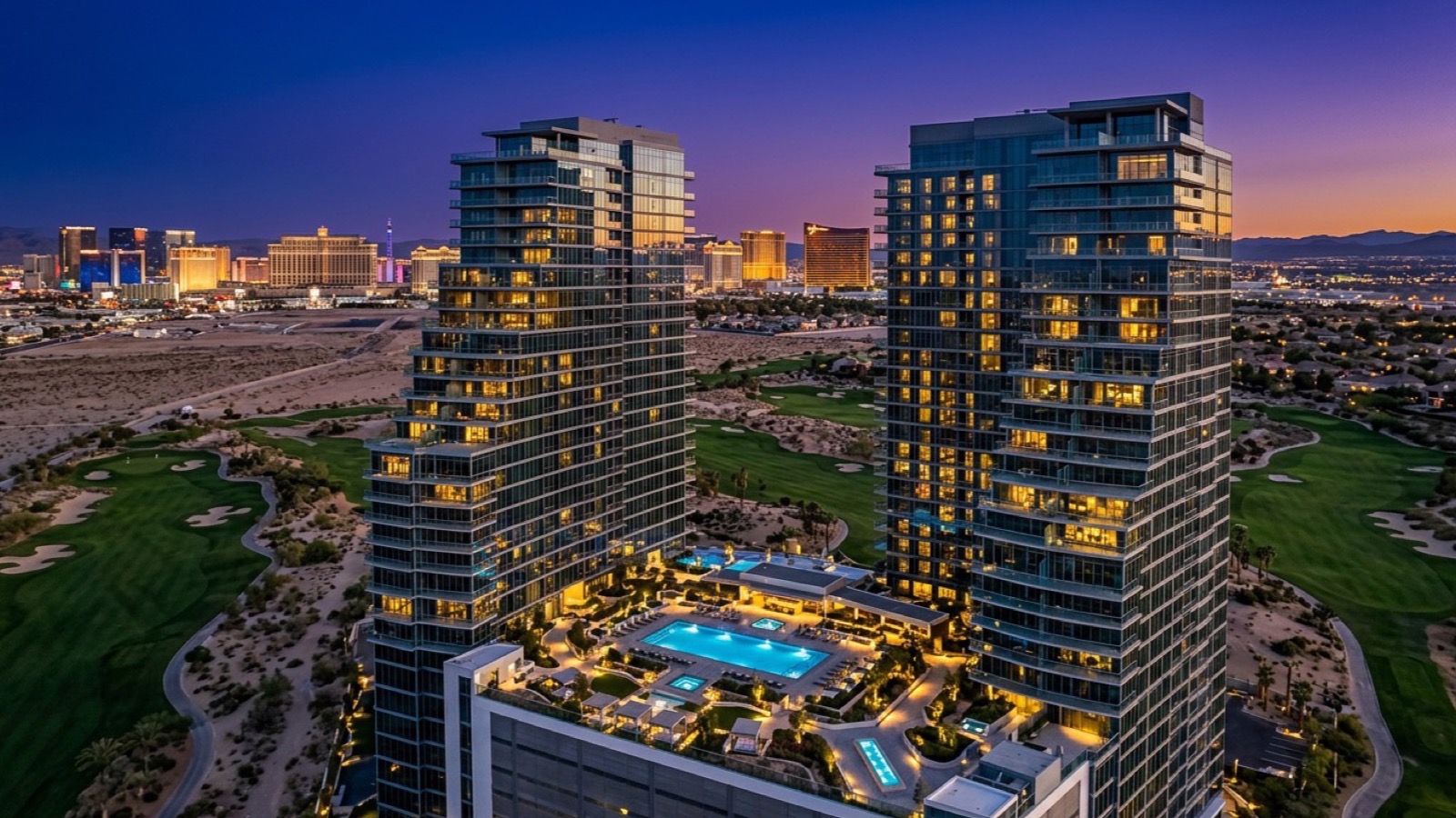 Twin residential condo towers beside a golf course at twilight with the Las Vegas Strip skyline in the distance