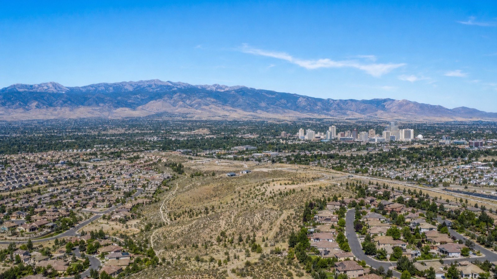 Aerial view across suburban neighborhoods and open sagebrush toward the downtown Reno skyline and distant mountains