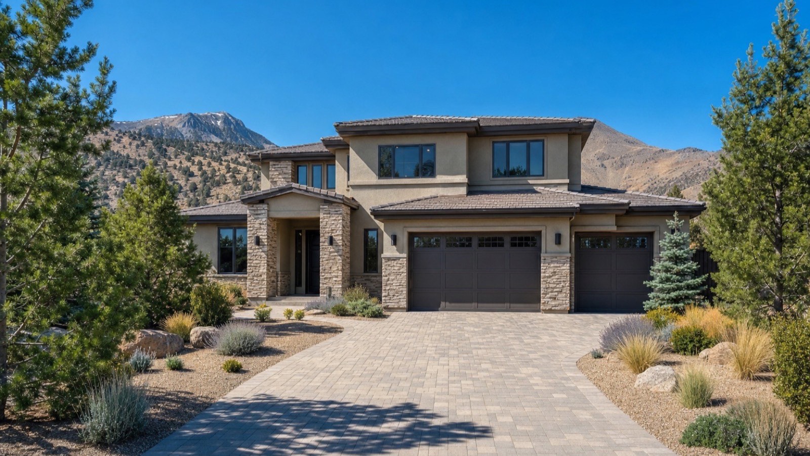 Two-story stone and stucco home with a wide paver driveway, pine trees and a high-desert peak behind