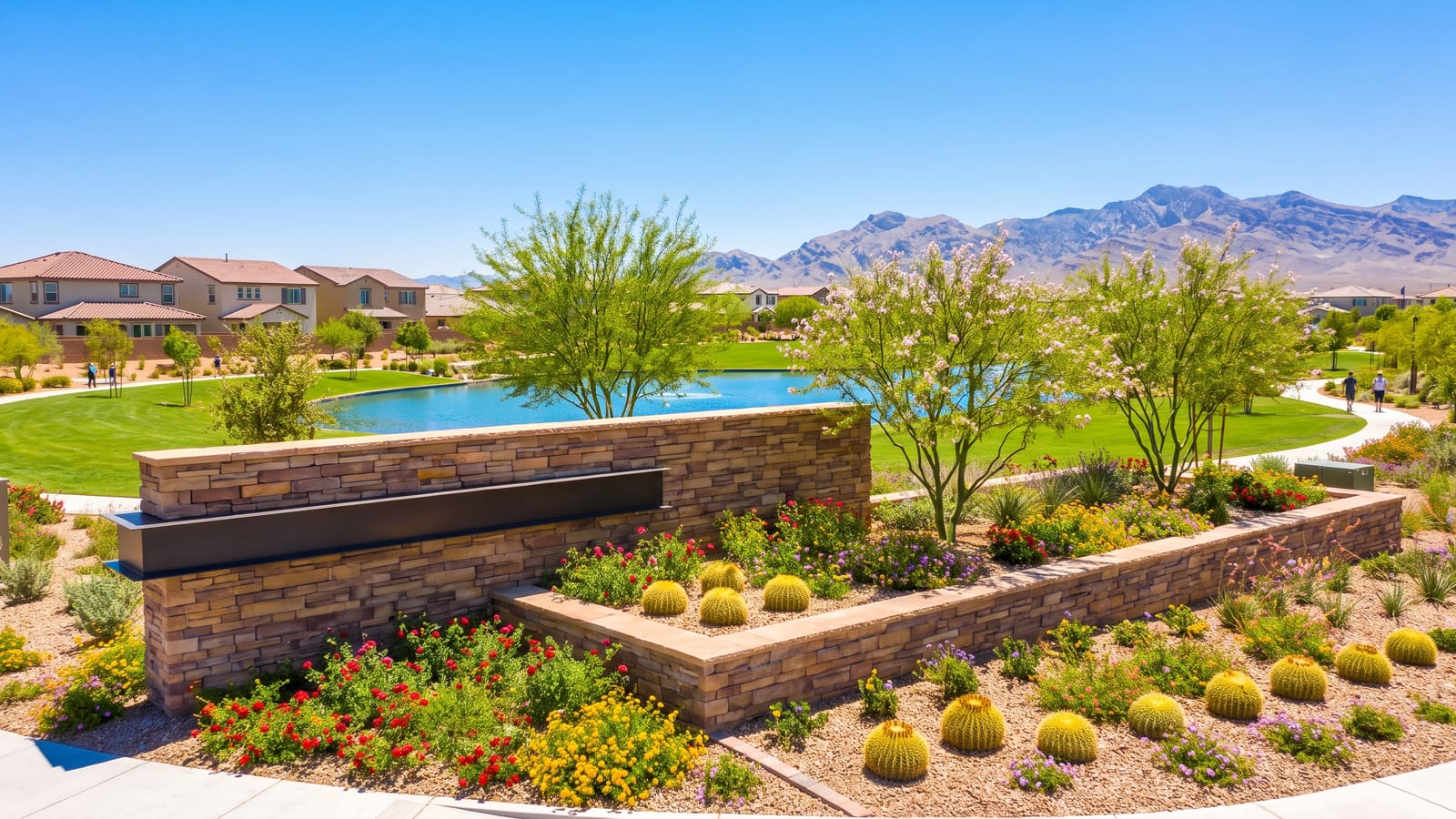 New North Las Vegas master-planned community entry monument with stone signage, blooming desert landscaping, a park lake, new homes, and the Sheep Range mountains